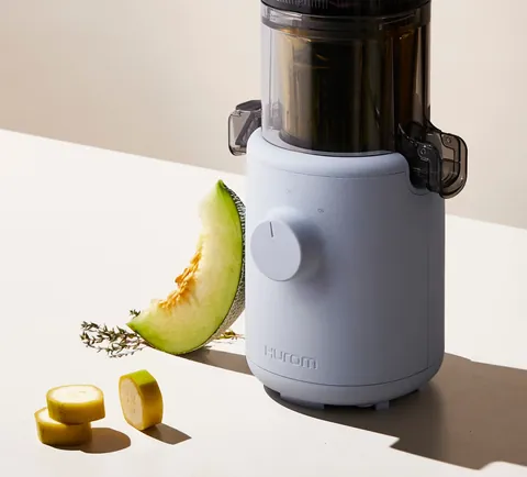 Błękitna wyciskarka Hurom H310A z bliska obok kawałka melona i plastrów banana