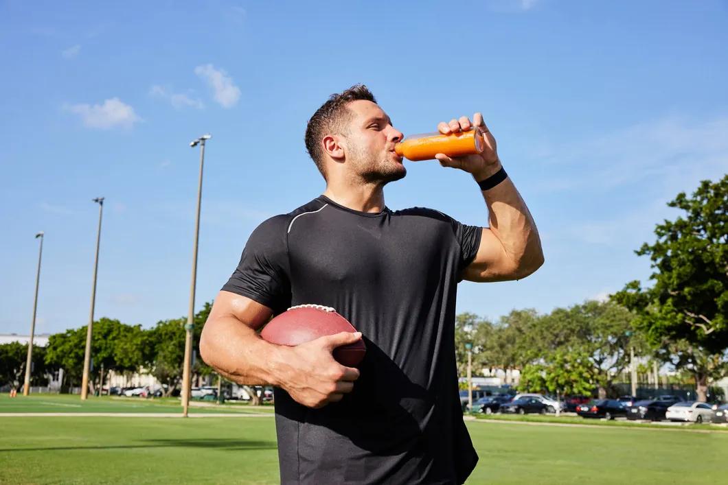 Nick Bosa pije sok z butelki, w drugiej ręce trzyma piłkę do futbolu amerykańskiego