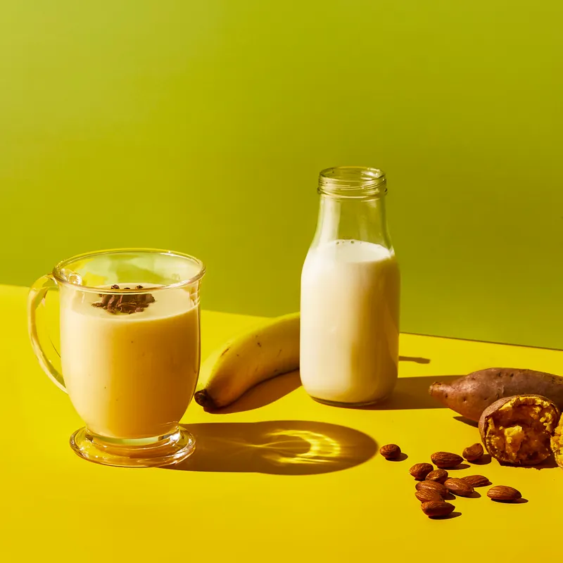 Koktajl bananowy w szklanym kubku, butelka mleka roślinnego, banan, migdały i batat na żółtym tle